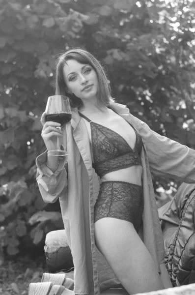 かなり若いですブルネット女性身に着けていますでダークランジェリー開催ガラスのワインとポージングオン革ソファ近く木製の家屋外 — ストック写真