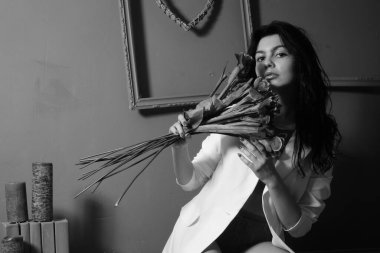 İç çamaşırlı ve ceketli güzel seksi bir kız elinde çiçeklerle stüdyoda kameraya poz veriyor, siyah beyaz bir fotoğraf.