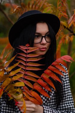 Siyah şapkalı genç bir kız kameraya sonbahar yapraklarıyla poz veriyor.