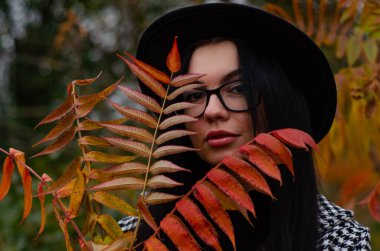 Siyah şapkalı genç bir kız kameraya sonbahar yapraklarıyla poz veriyor.