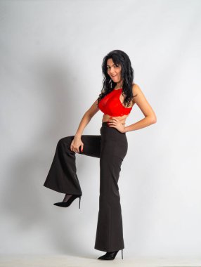 Kırmızı bluz ve siyah pantolon giyen çekici, genç, esmer manken beyaz odadaki kameraya poz veriyor.