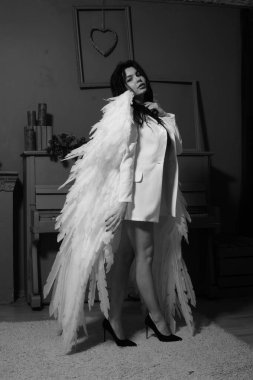 İç çamaşırlı, ceketli ve kanatlı güzel bir kız stüdyoda kameraya poz veriyor, siyah beyaz bir fotoğraf.