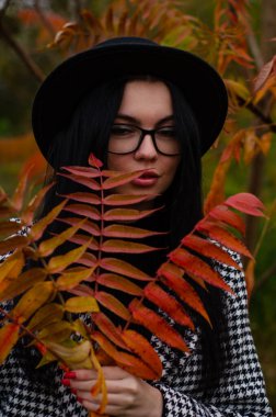 Siyah şapkalı genç bir kız kameraya sonbahar yapraklarıyla poz veriyor.