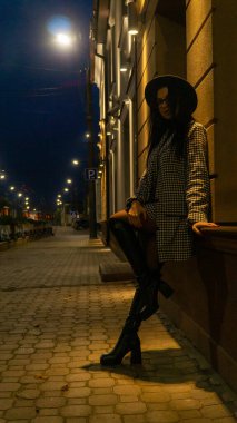 Siyah şapkalı genç ve güzel bir kız gece şehirde yürüyor ve kameraya poz veriyor.