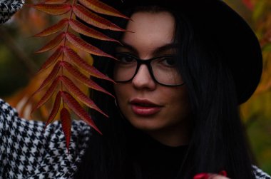 Siyah şapkalı genç bir kız kameraya sonbahar yapraklarıyla poz veriyor.