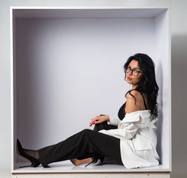 Beyaz odadaki kameraya poz veren siyah bluzlu ve beyaz ceketli çekici genç esmer manken.