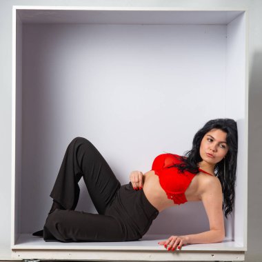 Kırmızı bluz ve siyah pantolon giyen çekici, genç, esmer manken beyaz odadaki kameraya poz veriyor.