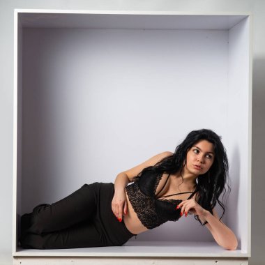 Siyah bluz ve siyah pantolon giyen çekici, genç, esmer manken beyaz odadaki kameraya poz veriyor.