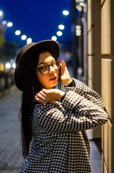 Siyah şapkalı genç ve güzel bir kız gece şehirde yürüyor ve kameraya poz veriyor.