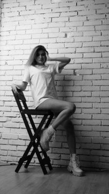 Stüdyoda sandalyenin yanında uzun saçlı, siyah beyaz bir kadın poz veriyor..  