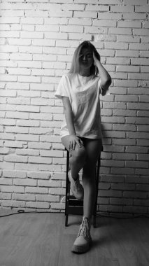 Uzun saçlı, uzun tişörtlü, stüdyoda sandalyeyle poz veren güzel bir kadının siyah beyaz fotoğrafı..  