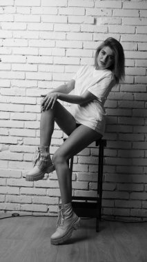 Uzun saçlı, uzun tişörtlü, stüdyoda sandalyeyle poz veren güzel bir kadının siyah beyaz fotoğrafı..  