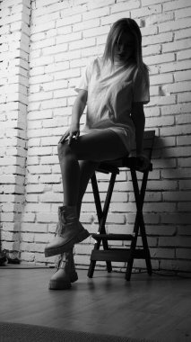 Uzun saçlı, uzun tişörtlü, stüdyoda sandalyeyle poz veren güzel bir kadının siyah beyaz fotoğrafı..  