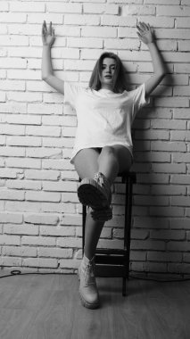 Uzun saçlı, uzun tişörtlü, stüdyoda sandalyeyle poz veren güzel bir kadının siyah beyaz fotoğrafı..  