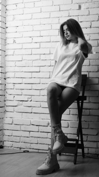 Uzun saçlı, uzun tişörtlü, stüdyoda sandalyeyle poz veren güzel bir kadının siyah beyaz fotoğrafı..  