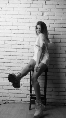 Uzun saçlı, uzun tişörtlü, stüdyoda sandalyeyle poz veren güzel bir kadının siyah beyaz fotoğrafı..  