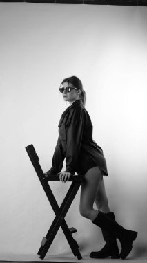 Siyah gömlekli ve güneş gözlüklü genç bir kadının stüdyoda poz verirken çekilmiş siyah beyaz bir fotoğrafı..  