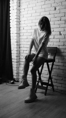 Uzun saçlı, uzun tişörtlü, stüdyoda sandalyeyle poz veren güzel bir kadının siyah beyaz fotoğrafı..  