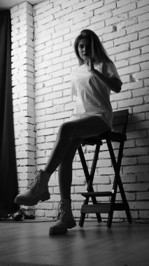Uzun saçlı, uzun tişörtlü, stüdyoda sandalyeyle poz veren güzel bir kadının siyah beyaz fotoğrafı..  