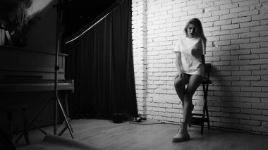 Uzun saçlı, uzun tişörtlü, stüdyoda sandalyeyle poz veren güzel bir kadının siyah beyaz fotoğrafı..  