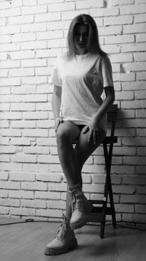 Uzun saçlı, uzun tişörtlü, stüdyoda sandalyeyle poz veren güzel bir kadının siyah beyaz fotoğrafı..  