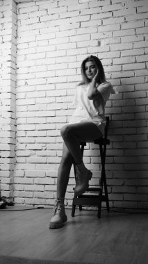 Uzun saçlı, uzun tişörtlü, stüdyoda sandalyeyle poz veren güzel bir kadının siyah beyaz fotoğrafı..  