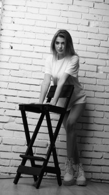 Stüdyoda sandalyenin yanında uzun saçlı, siyah beyaz bir kadın poz veriyor..  