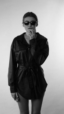 Siyah gömlekli ve güneş gözlüklü genç bir kadının stüdyoda poz verirken çekilmiş siyah beyaz bir fotoğrafı..  