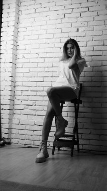 Uzun saçlı, uzun tişörtlü, stüdyoda sandalyeyle poz veren güzel bir kadının siyah beyaz fotoğrafı..  