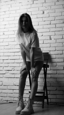 Uzun saçlı, uzun tişörtlü, stüdyoda sandalyeyle poz veren güzel bir kadının siyah beyaz fotoğrafı..  