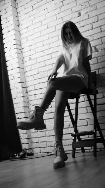 Uzun saçlı, uzun tişörtlü, stüdyoda sandalyeyle poz veren güzel bir kadının siyah beyaz fotoğrafı..  