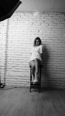 Uzun saçlı, uzun tişörtlü, stüdyoda sandalyeyle poz veren güzel bir kadının siyah beyaz fotoğrafı..  