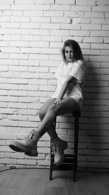 Uzun saçlı, uzun tişörtlü, stüdyoda sandalyeyle poz veren güzel bir kadının siyah beyaz fotoğrafı..  