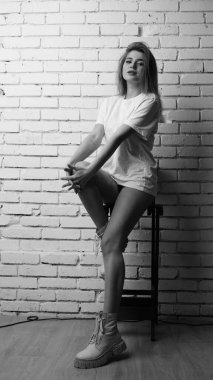 Uzun saçlı, uzun tişörtlü, stüdyoda sandalyeyle poz veren güzel bir kadının siyah beyaz fotoğrafı..  