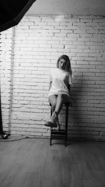 Uzun saçlı, uzun tişörtlü, stüdyoda sandalyeyle poz veren güzel bir kadının siyah beyaz fotoğrafı..  