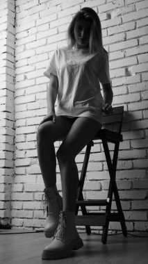 Uzun saçlı, uzun tişörtlü, stüdyoda sandalyeyle poz veren güzel bir kadının siyah beyaz fotoğrafı..  