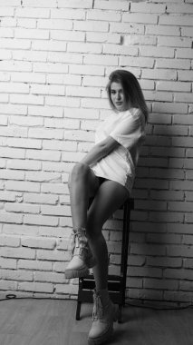 Uzun saçlı, uzun tişörtlü, stüdyoda sandalyeyle poz veren güzel bir kadının siyah beyaz fotoğrafı..  