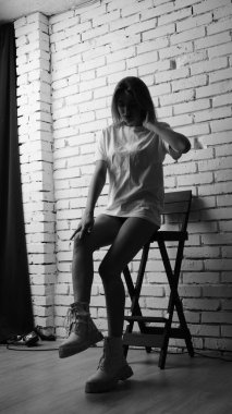 Uzun saçlı, uzun tişörtlü, stüdyoda sandalyeyle poz veren güzel bir kadının siyah beyaz fotoğrafı..  