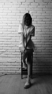 Uzun saçlı, uzun tişörtlü, stüdyoda sandalyeyle poz veren güzel bir kadının siyah beyaz fotoğrafı..  