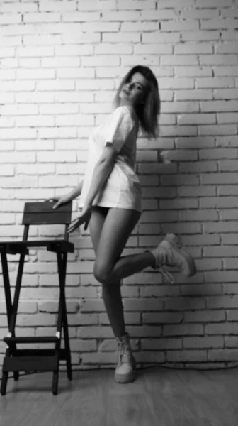身穿T恤衫 留着长发的年轻漂亮女子站在演播室椅子旁边的黑白照片 — 图库照片