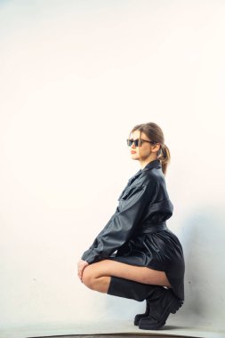 Uzun saçlı, siyah gömlekli ve güneş gözlüklü güzel, şehvetli bir kadının fotoğrafı stüdyoda beyaz bir duvara yaslanmış..  