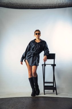 Uzun saçlı, siyah gömlekli ve güneş gözlüklü güzel, şehvetli bir kadının fotoğrafı stüdyoda beyaz bir duvara yaslanmış..  