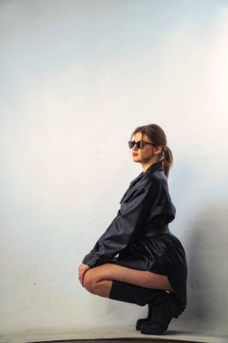 Uzun saçlı, siyah gömlekli ve güneş gözlüklü güzel, şehvetli bir kadının fotoğrafı stüdyoda beyaz bir duvara yaslanmış..  