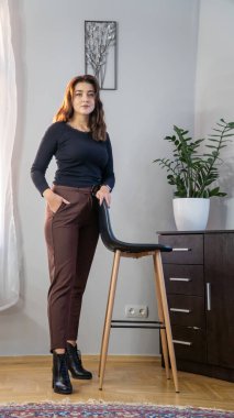 Güzel, genç, esmer kadın. Günlük giysiler içinde. Odada kameraya poz veriyor.