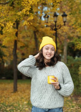 Sonbahar parkında elinde fincanı tutan sarı şapkalı güzel bir kadın. Genç bayan dışarıda sıcak kahve içiyor. Sonbahar mevsimi konsepti.