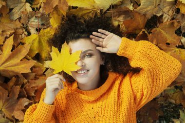 Sonbahar yaprağıyla gülümseyen genç kadın sonbahar parkında eğleniyor. Kıvırcık saçlı mutlu bir kadının portresi açık havadaki yeşillikte yatıyor..