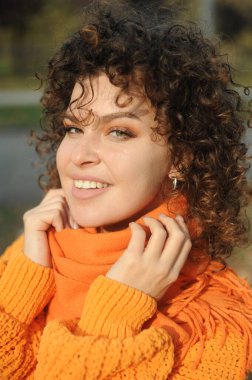 Sonbahar parkında turuncu kazaklı, gülümseyen kıvırcık saçlı kadın..