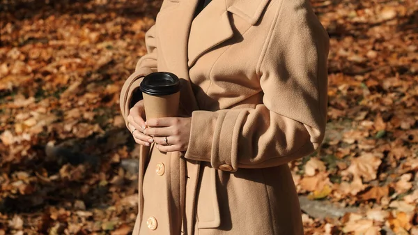 コーヒー屋外のカップで流行のベージュのコートで若い巻き毛の女性を笑顔 紅葉の美しい秋の公園でコーヒーを飲む若い女性 — ストック写真