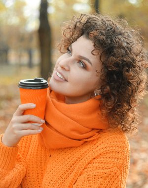 Gülümseyen kıvırcık saçlı turuncu kazaklı güzel bir kadın cep telefonundan konuşuyor. 