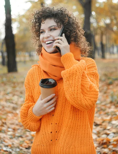 주황색 스웨터를 핸드폰으로 말하는 곱슬곱슬 머리의 아름다운 — 스톡 사진